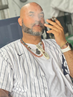 Picture of Juan Gonzalez Senior wearing a white shirt with black stripes and a necklace, he hs a close shaved beard and is bald. Juan is smoking a cigar and the smoke is around his face.